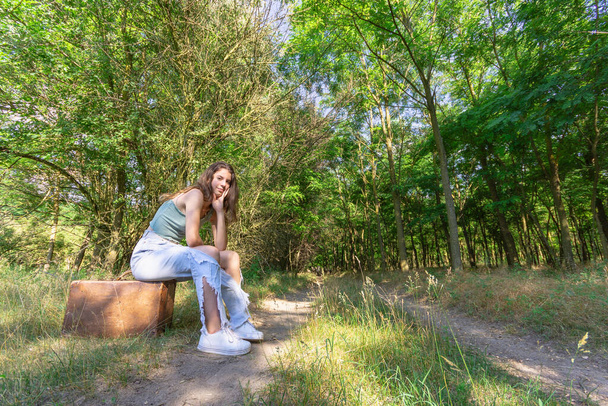 Młoda kobieta siedzi na starej brązowej walizce na leśnej drodze gruntowej - Zdjęcie, obraz