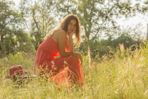 Jonge vrouw in rode jurk zittend op koffer en kijkend in de camera in het bos met achtergrondverlichting - Foto, afbeelding