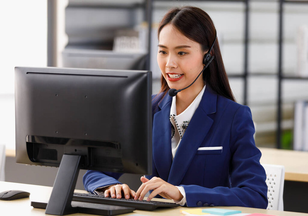 Mladý dlouhý černý vlasy šťastný jistý asijské žena operátor nosit modrý formální oblek mluvit přes černý sluchátka set pracovní poradenství pomáhá zákazníkovi pomocí počítače monitor sám v kanceláři místnosti. - Fotografie, Obrázek