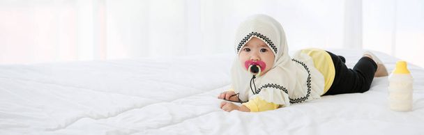 Ein niedliches muslimisches Baby im Hijab-Kleid mit Schnuller in der Hand krabbelt auf dem Bett in der Wohnung. - Foto, Bild