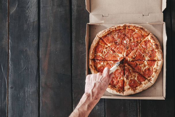 Male hand cuts Pepperoni pizza with a round knife on a dark wooden background. Delicious and fresh pizza in a box with round pieces of sausage, on a wooden background. Pizza delivery - Photo, Image