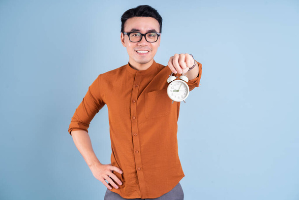 Joven asiático hombre celebración reloj en azul fondo - Foto, imagen