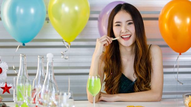 Asian beautiful girl sit smile holding eating french fries in hand at beverage table with alcohol drinks bottles tall wine glasses and cookies in front colorful balloons background at Christmas party. - Photo, Image
