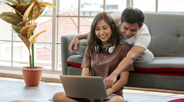 Young attractive Asian couple sitting and looking at laptop together in living room. Woman sitting on the floor wearing headphones and man lying on couch. Concept for love and couple relationship. - Photo, Image