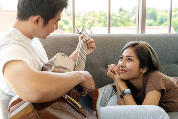 Giovane attraente coppia asiatica insieme su un divano. Uomo seduto a suonare la chitarra e donna sdraiato e in ascolto in soggiorno con gli occhiali di sfondo della finestra. Concetto di amore e relazione di coppia. - Foto, immagini