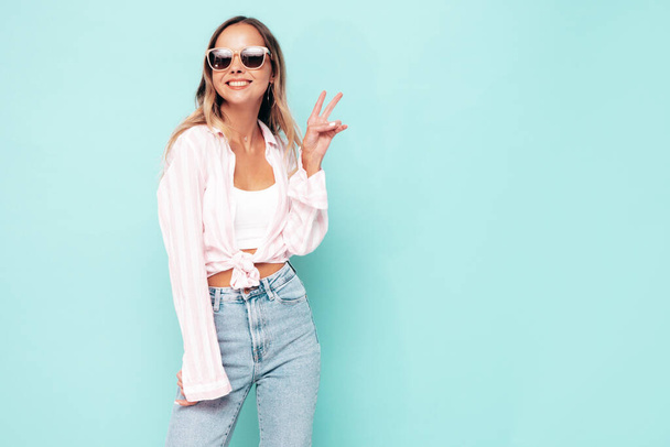 Young beautiful smiling female in trendy summer clothes. Sexy carefree woman posing near blue wall in studio. Positive model having fun indoors. Cheerful and happy. Shows peace sign, in sunglasses - Zdjęcie, obraz