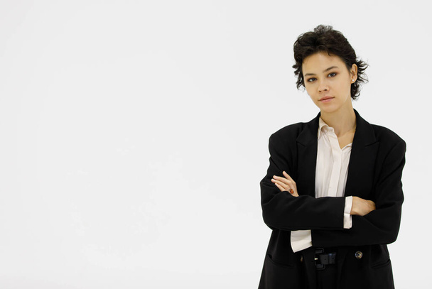 Confident well dressed female entrepreneur in black formal suit standing with crossed arms on white background in studio and looking at camera - Foto, afbeelding
