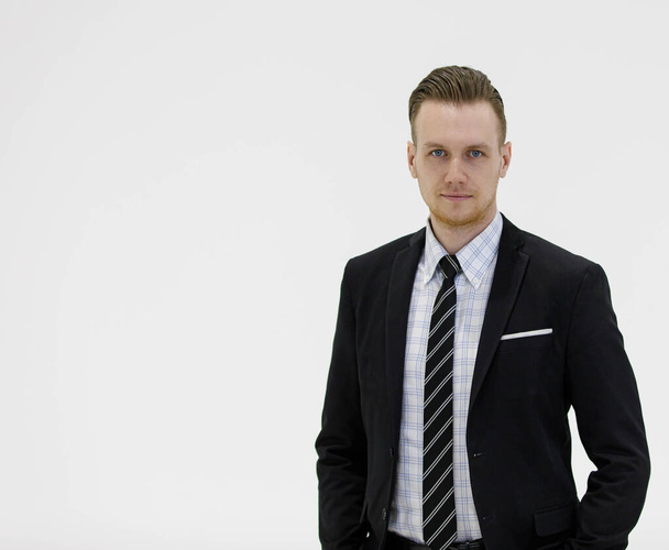Самоуверенный предприниматель в элегантном черном костюме смотрит в камеру, стоя с руками в карманах на белом фоне в студии - Фото, изображение