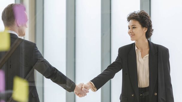 Side view of glad male and female business partners shaking hands after successful deal and looking at each other while standing in modern workspace - Photo, Image