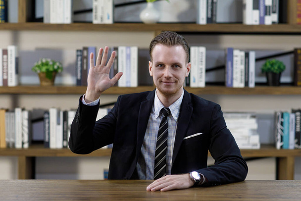 Amable empresario masculino en traje sentado en la mesa y saludando la mano mientras muestra gesto de saludo y mirando a la cámara contra estante en el lugar de trabajo - Foto, Imagen