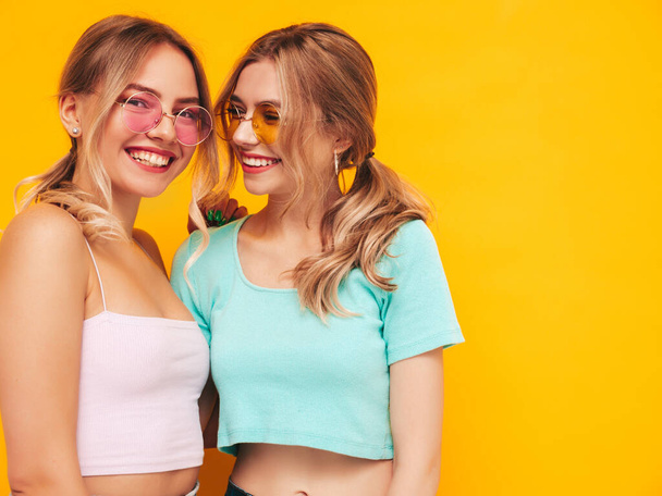 Two young beautiful smiling blond hipster female in trendy summer clothes. Sexy carefree women posing near yellow wall in studio. Positive models having fun. Cheerful and happy. In sunglasses - Фото, изображение