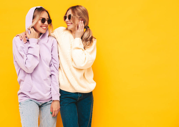 Dvě mladé krásné usměvavé blonďaté hipsterky v módních letních šatech. Sexy bezstarostné ženy pózující u žluté zdi ve studiu. Pozitivní modely se baví. Veselý a šťastný. In sunglasses - Fotografie, Obrázek