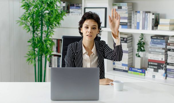 Alegre mujer empresaria saludando de la mano y mostrando gesto de saludo mientras se sienta en la mesa con el ordenador portátil en la oficina y mirando a la cámara - Foto, imagen