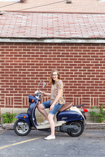 PRO, UNITED STATES - Jun 21, 2019: A vertical shallow focus shot of a smiling girl with glasses on motor scooter in front of a brick house in the city of Provo in Utah, United States - Valokuva, kuva