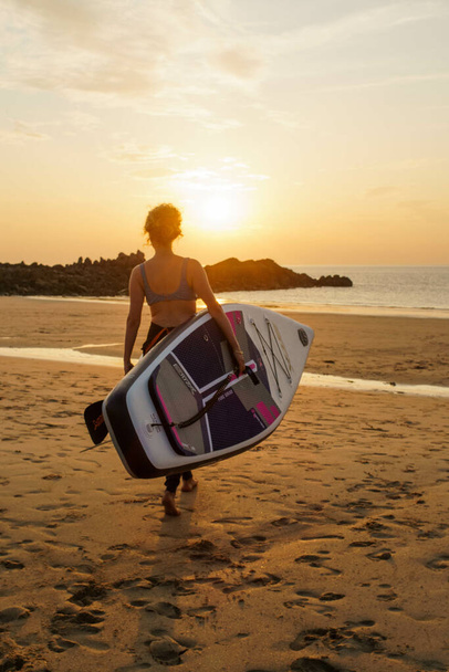 NORMANDY, FRANCE - Aug 06, 2021: A Caucasian girl with SUP board and paddle walking towards the ocean during a golden sunset in summer - Foto, Imagen
