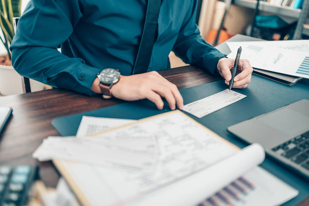 Business man hand writing and signing white blank bank check book and dollar bill, coin, laptop and graph chart on the desk at office. Payment by check, paycheck, payroll concept. - 写真・画像