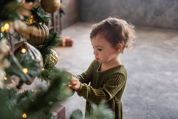 Little girl in warm green dress decorates the Christmas tree with elegant, stylish, golden balls, toys. Retro decor in gray loft interior. Home decoration for the holiday. Copy space, gray background - 写真・画像