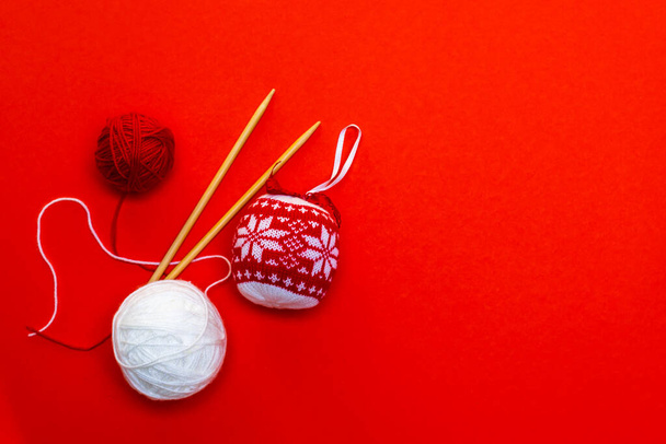 Gebreide Kerstbal en ballen van garen met breinaalden op een rode achtergrond. Kerstkaart, handwerk. Horizontale foto. - Foto, afbeelding