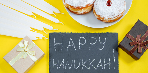 Feliz Hanukkah. Postre judío Sufganiyot sobre fondo amarillo. Símbolos de la fiesta del judaísmo religioso. Donuts, velas y regalos. Formato de banner - Foto, imagen