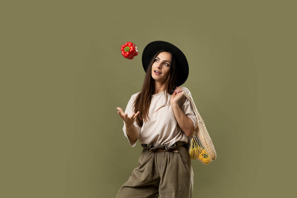 Smiling brunette young woman in a beige t-shirt and a hat holding a reusable mesh bag with fresh vegetables and fruits on a shoulder and throwing a red pepper in a air. Zero waste concept. - Photo, image