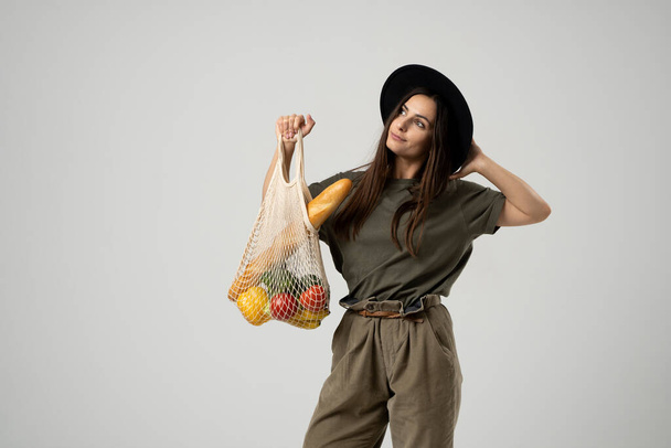 A young fashion brunete girl in a beige t-shirt and black hat holds a mesh reusable eco bag with food, vegetabes, fruits. Zero waste, plastic free. Eco friendly concept. Sustainable lifestyle. - Foto, imagen