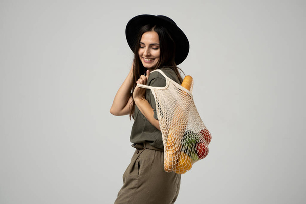 Zero waste concept. A young girl holds on her shoulder a textile mesh eco bag with a groceries. The girl smiles, wearing a beige t-shirt and hat. Refusal of plastic bags. - Foto, afbeelding