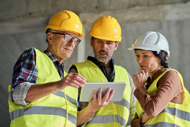 Tres ingenieros caucásicos discutiendo sobre tableta digital en el sitio de construcción - Foto, Imagen