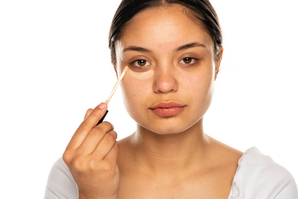 closeup μιας νεαρής όμορφης γυναίκας εφαρμόζει concealer κάτω από το μάτι της σε λευκό φόντο - Φωτογραφία, εικόνα