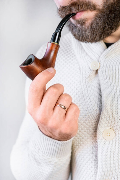 Mężczyzna dżentelmen z brodą i wąsami w jasnym kardiganie trzyma w ręku i pali fajkę, elegancki modny człowiek - Zdjęcie, obraz