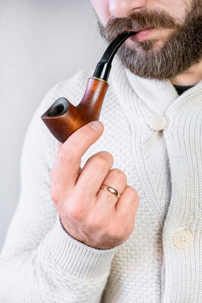Mężczyzna dżentelmen z brodą i wąsami w jasnym kardiganie trzyma w ręku i pali fajkę, elegancki modny człowiek - Zdjęcie, obraz