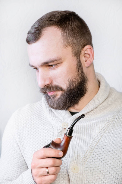 Улыбающийся джентльмен с бородой и усами в легком кардигане держит и курит трубку, стильный модный модный мужчина - Фото, изображение
