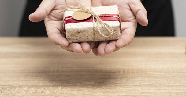 Коричневая подарочная коробка и красная лента на руках над коричневым столом для подарка тем, кого ты любишь - Фото, изображение