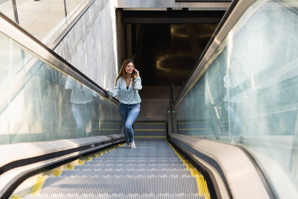 Привлекательная деловая женщина в очках с помощью смартфона, улыбаясь, поднимается на метро, метро, эскалатор аэропорта. Концепция бизнеса - Фото, изображение