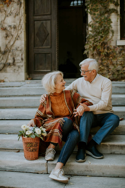 Όμορφο ζευγάρι ηλικιωμένων κάθεται στις σκάλες με καλάθι γεμάτο λουλούδια και παντοπωλεία - Φωτογραφία, εικόνα