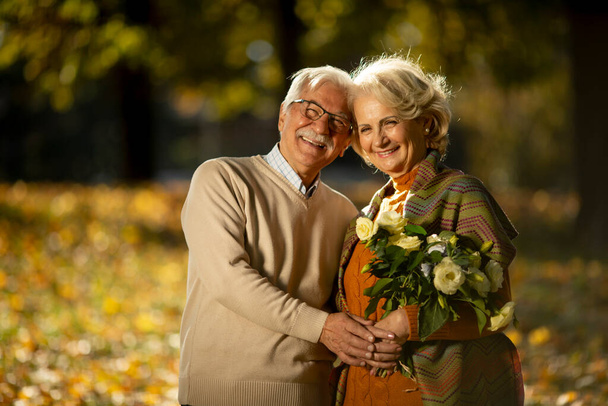 Hermosa pareja de ancianos abrazando con ramo de flores en el parque de otoño - Foto, imagen