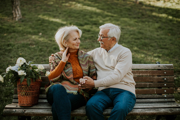 Красивая пожилая пара сидит на скамейке с корзиной, полной цветов и обнимает - Фото, изображение