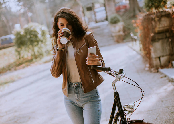 Pretty young woman with mobile phone drink coffee to go by the bicycle on autumn day - Foto, afbeelding