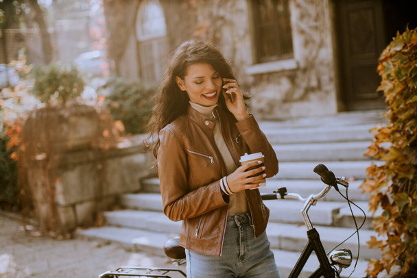 Pretty young woman with mobile phone drink coffee to go by the bicycle on autumn day - Photo, Image