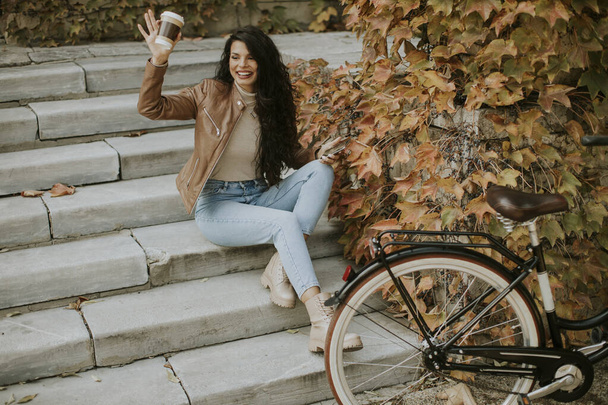 Pretty young woman with mobile phone drink coffee to go at the stairs by the bicycle on autumn day - Фото, изображение