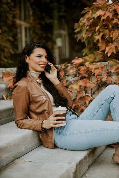 Pretty young woman with mobile phone drink coffee to go at the outdoor stairs on autumn day - Photo, Image