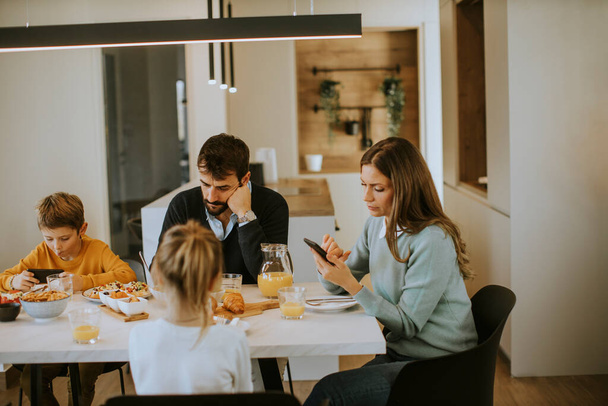 Семья с помощью мобильных телефонов во время завтрака за обеденным столом в квартире - Фото, изображение