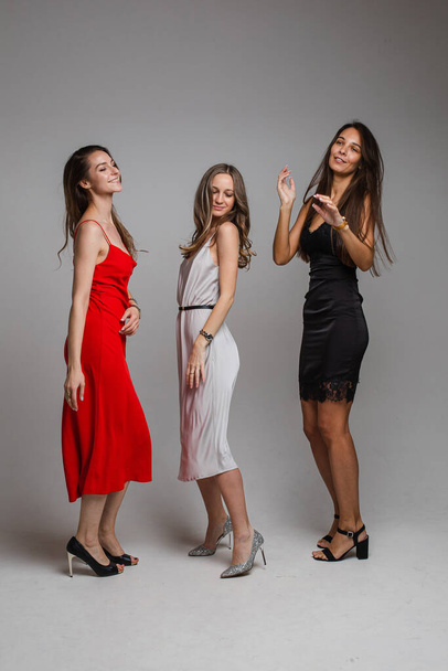 Charming positive young girls friends wearing stylish feminine evening dresses posing on gray studio background - Фото, изображение