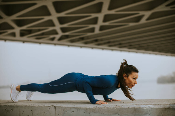 Young woman in blue track suit doing pushups under the bridge in the urban environment - Photo, Image