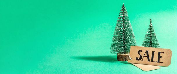 ターコイズを背景にしたクリスマスセールのコンセプト。装飾的なモミの木と紙のタグ伝統的な新年のシンボル。流行のハードライト、暗い影。フラットレイアウト、モックアップ、テンプレート、バナー形式 - 写真・画像