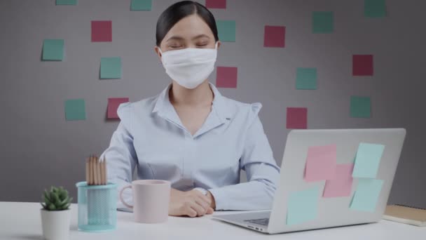 Asiatin trägt glücklich Maske und legt ihre Faust auf die linke Brust, um im Home Office Vertrauen zu zeigen. - Filmmaterial, Video