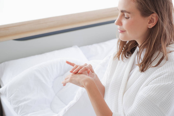 Красивая белая женщина наносит крем-увлажнитель на руку и сидит на кровати. - Фото, изображение