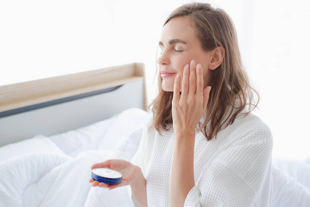 Mooie blanke vrouw aanbrengen moisturizer crème op gezicht en zitten op haar bed - Foto, afbeelding