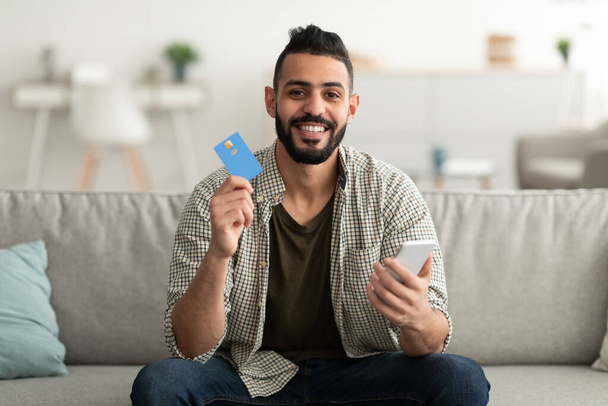 Objednávka zboží online. Šťastný mladý arabsky chlap s kreditní kartou a smartphone nakupování na webu doma - Fotografie, Obrázek