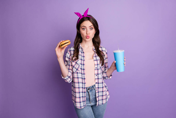 Foto retrato de menina bonita comendo hambúrguer bebendo refrigerante doce enviando beijo de ar isolado no fundo de cor púrpura brilhante - Foto, Imagem