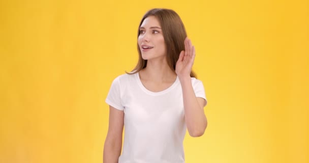 Jeune femme curieuse entendant des tintements autour, écoutant la caméra et de côté avec la main près de son oreille, fond jaune - Séquence, vidéo